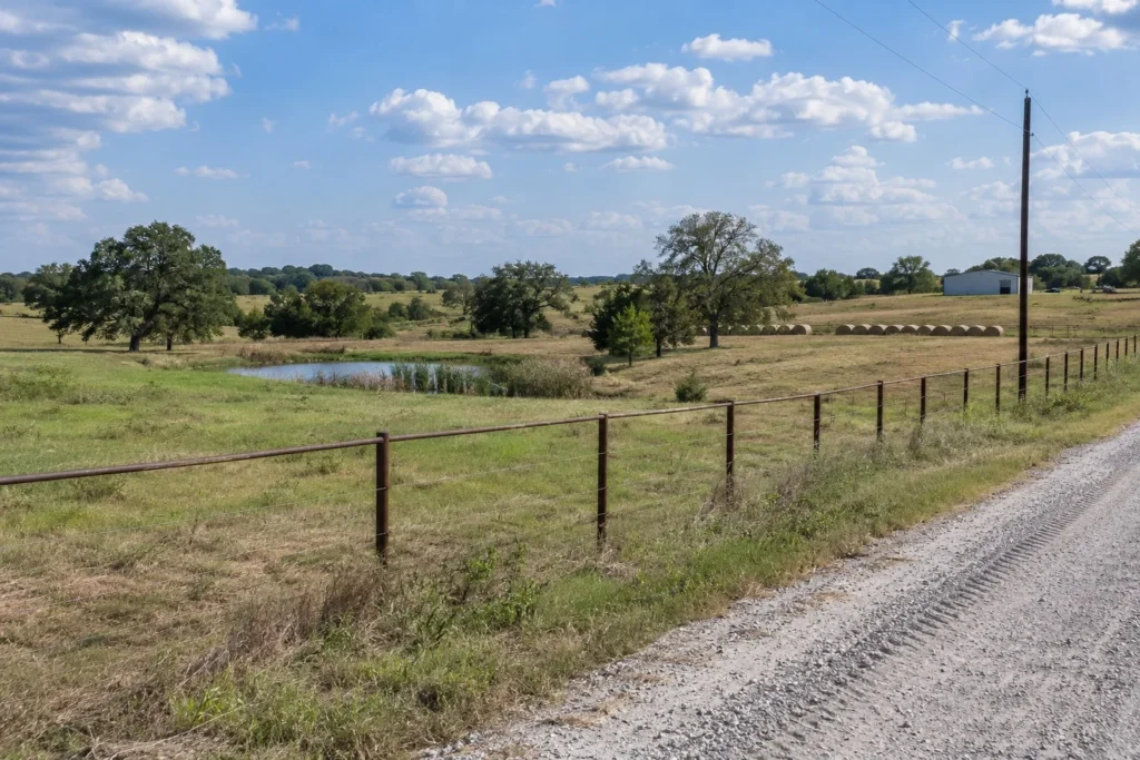 Rural homesite with a pipe-and-cable fence, stock pond, post oaks and round hay bales in North Texas